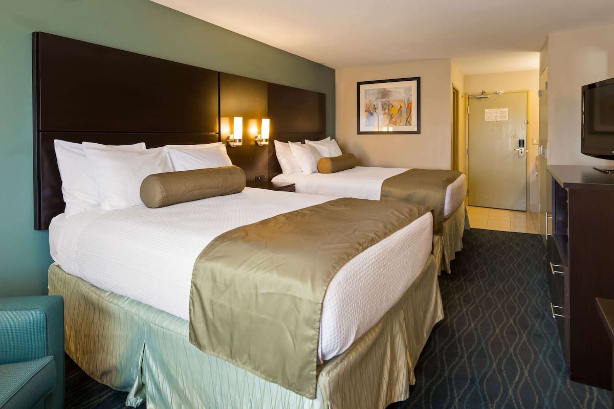 Quarto de hotel com duas camas, uma televisão e uma porta que dá para um corredor.