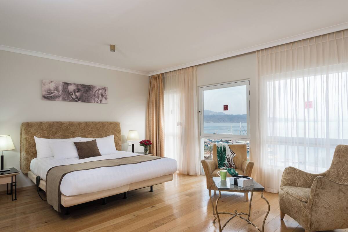 Een hotelkamer met een groot bed, een bureau met stoel, uitzicht op zee en een schilderij aan de muur.