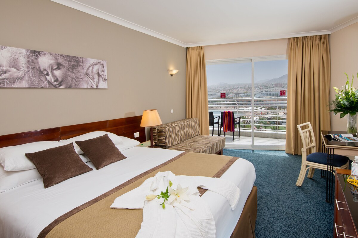 Une chambre d’hôtel avec un grand lit, un canapé, un bureau et une vue sur le paysage urbain depuis un balcon.