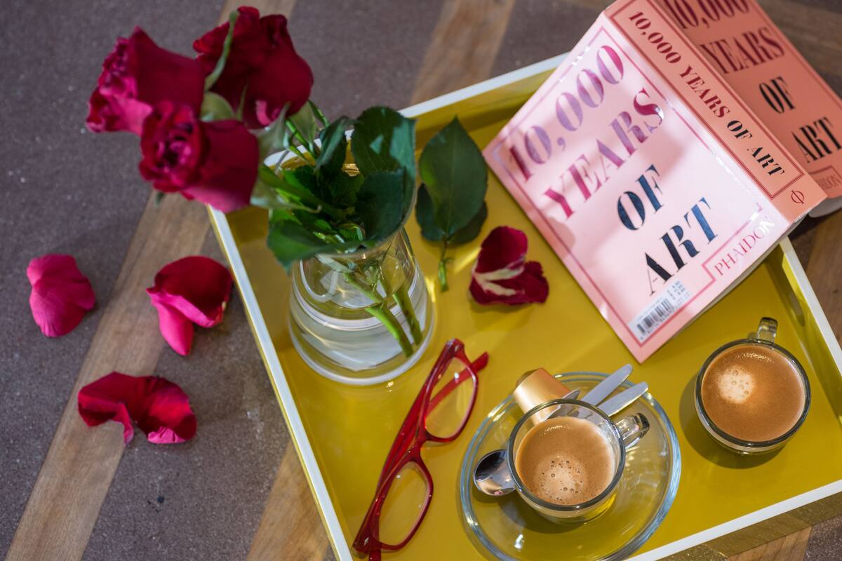 Een dienblad met een vaas rode rozen, een paar rode brillen, twee kopjes koffie en een boek getiteld '10.000 jaar kunst'.