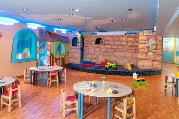 Zona de juegos infantiles con mesas, sillas y un escenario.