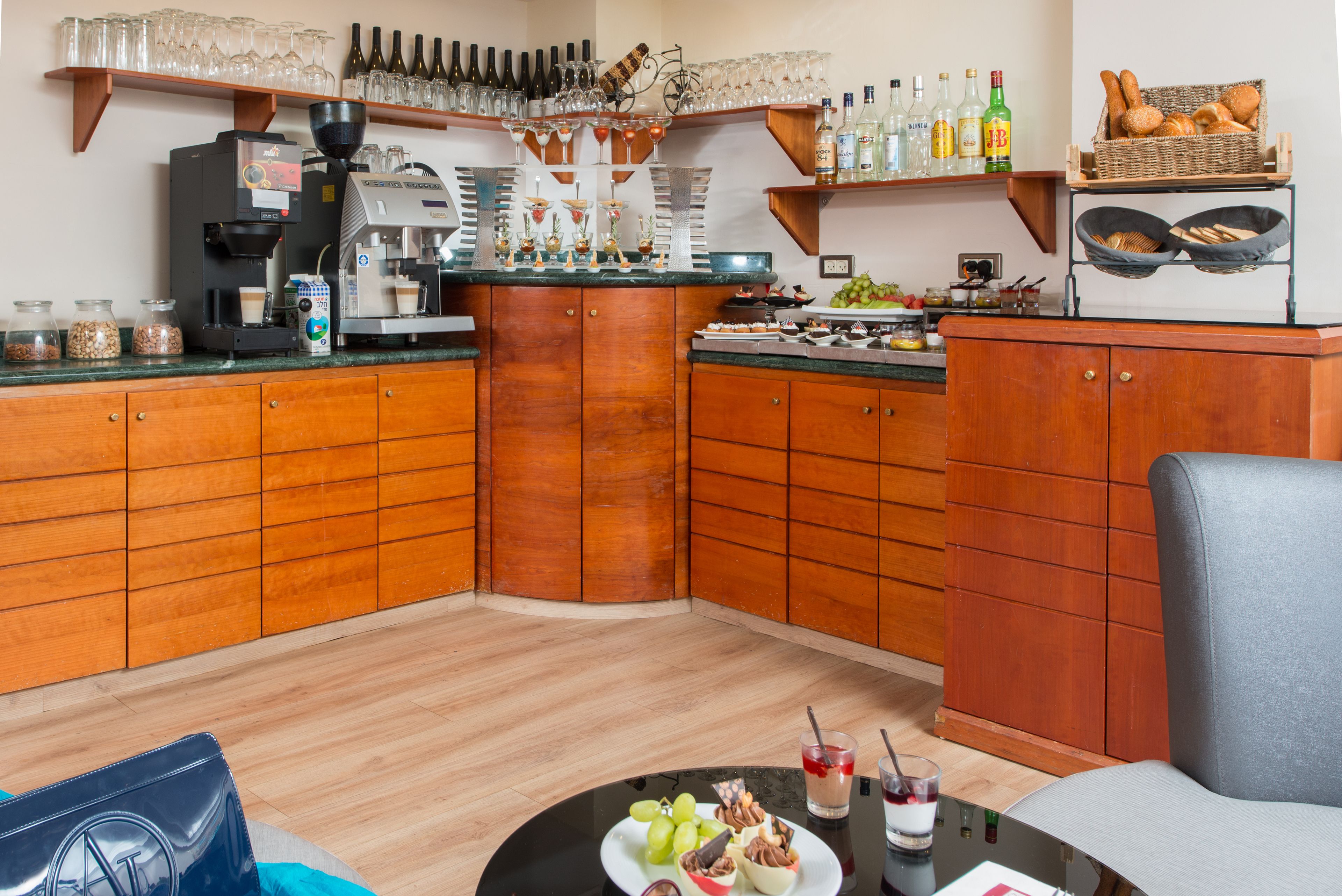 A corner bar area with a coffee machine, shelves with bottles and glasses, and a basket of bread.