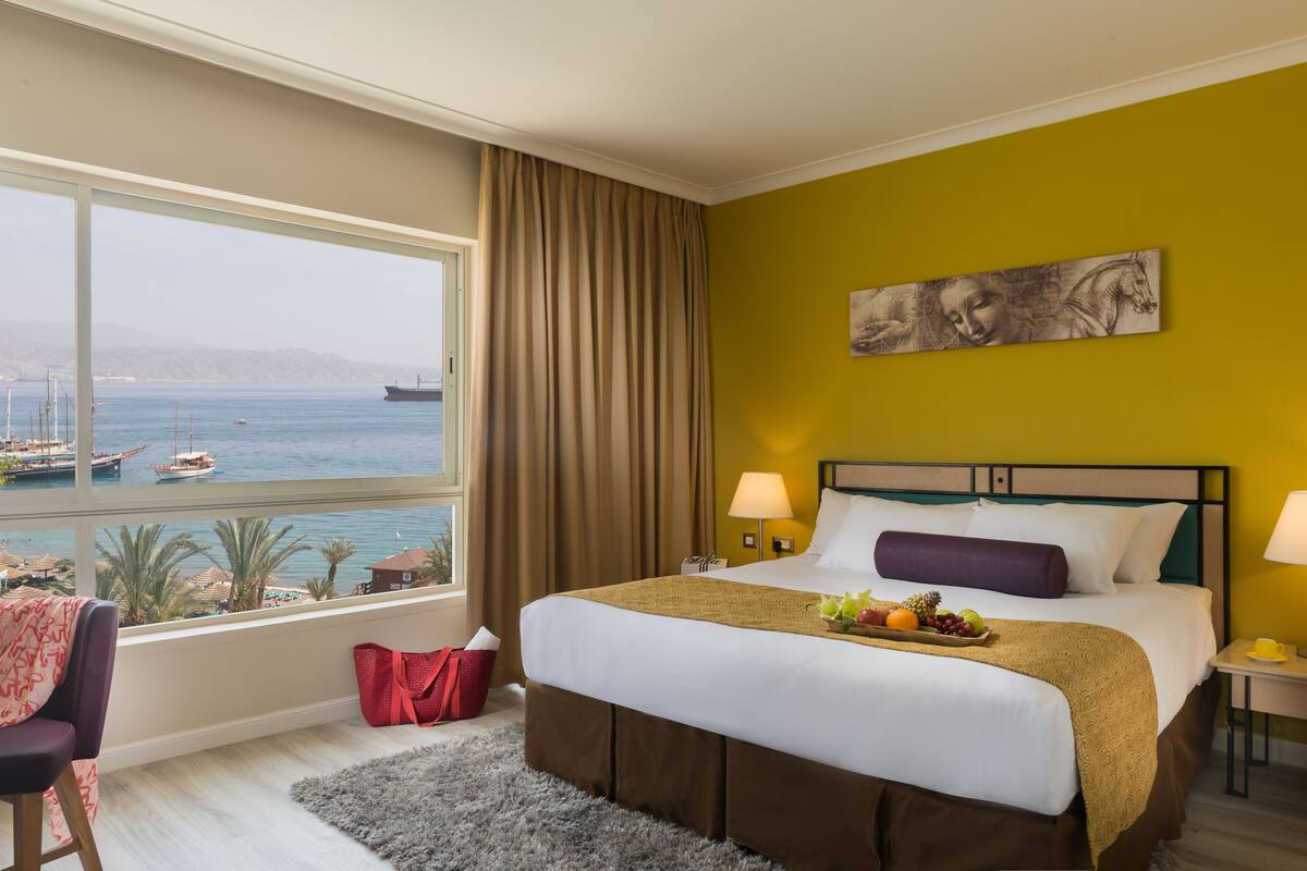 Een hotelkamer met een groot raam met uitzicht op zee en boten, een bed met een schaal fruit, en een schilderij aan de muur.