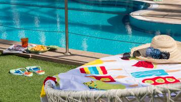 Zona de piscina con una silla de mimbre, una toalla colorida, chanclas, un sombrero, gafas de sol y un cuenco de fruta.