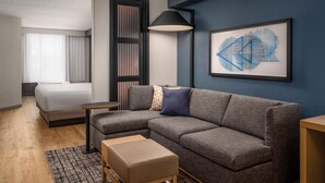 A modern hotel room with a gray sectional sofa, a small wooden table, a beige ottoman, a large abstract painting, and a bed with a white comforter.