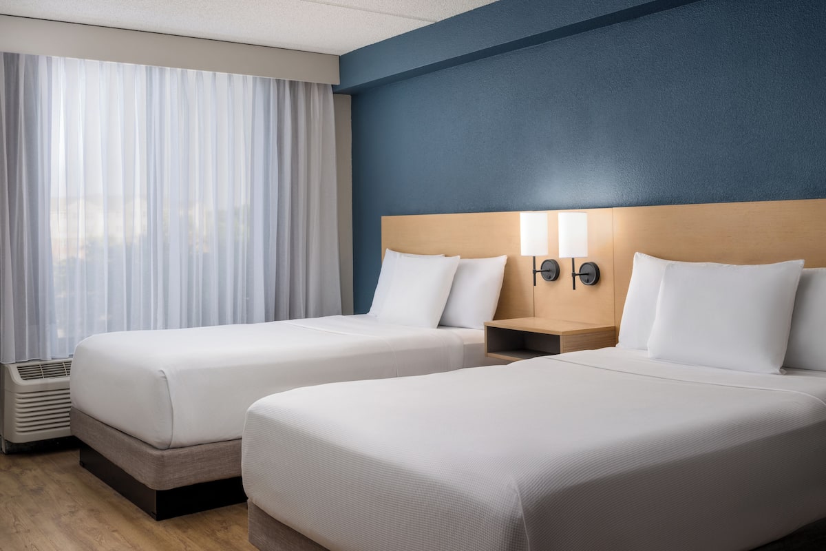 Ein Hotelzimmer mit zwei Betten, einem Fenster mit durchsichtigen Vorhängen, einer Wandlampe und einem Heizkörper.
