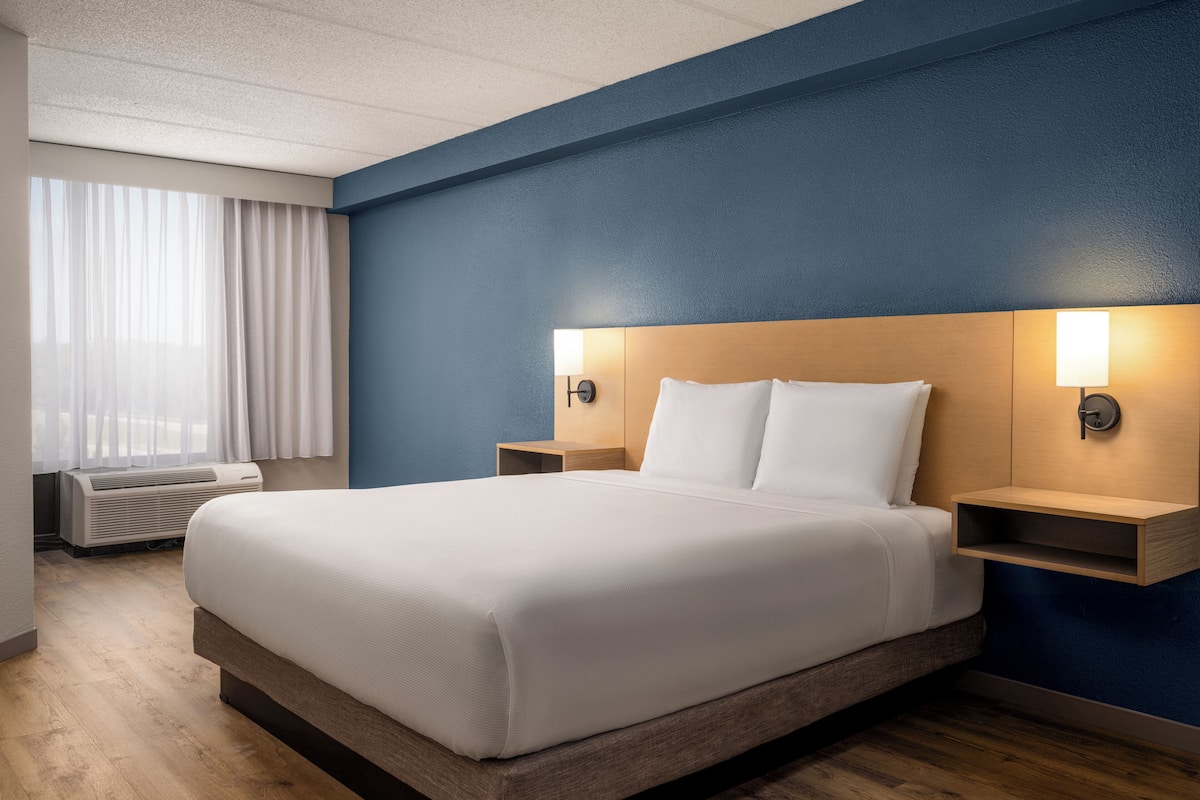 Ein Hotelzimmer mit einem großen Bett, einem Holzkopfende, zwei Nachttischen mit Lampen, einem Fenster mit Vorhängen und einer Klimaanlage.