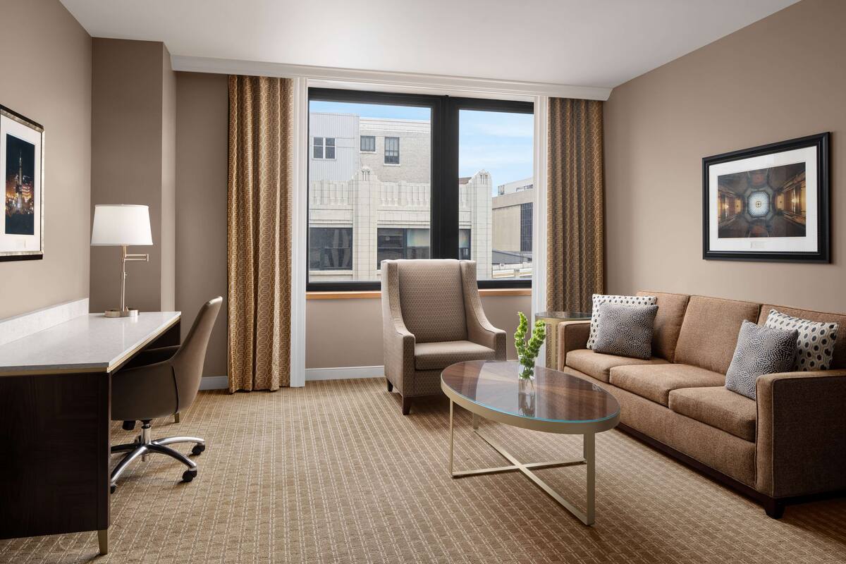 Een hotelkamer met een bureau, stoel, bank en een groot raam met gordijnen.