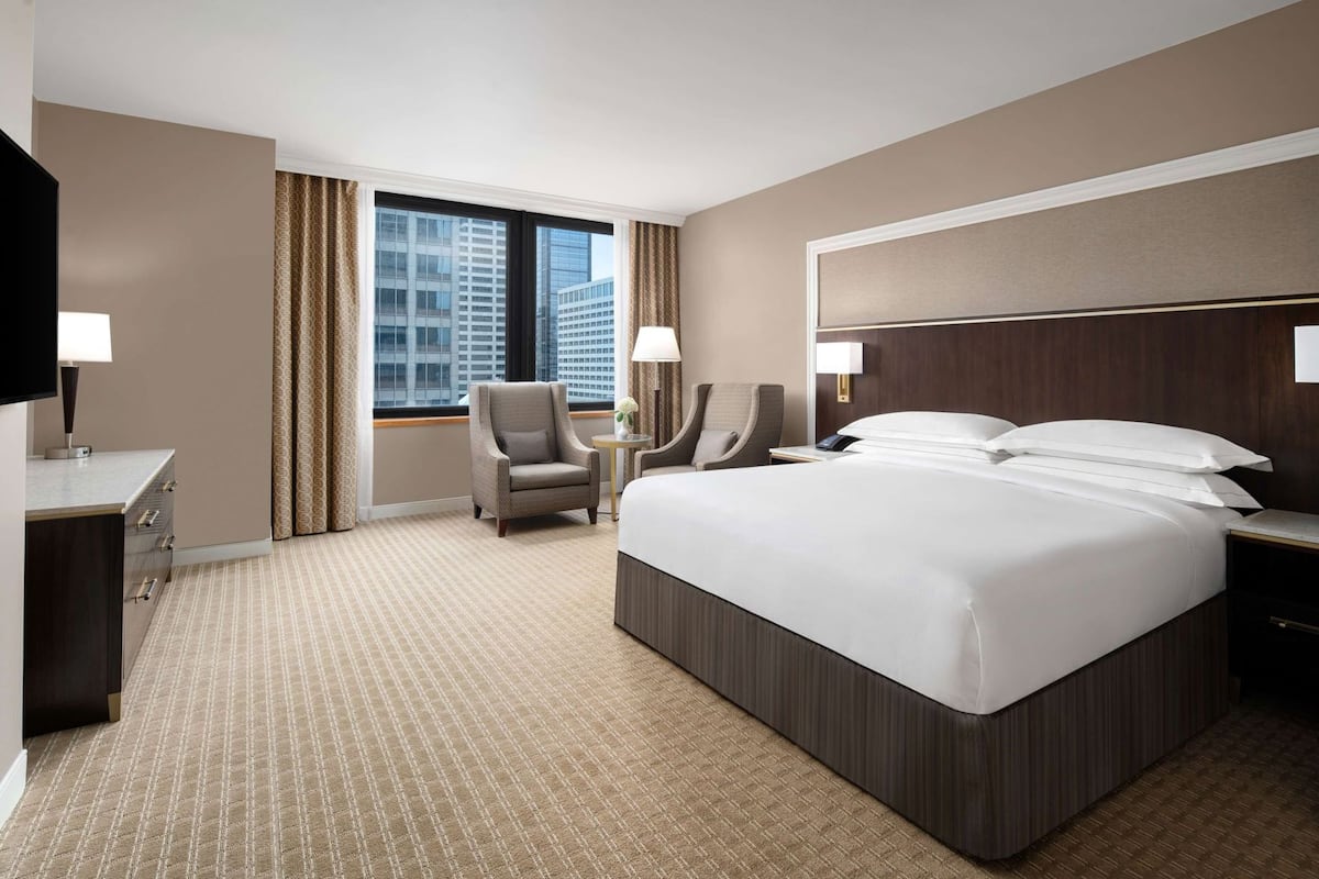Quarto de hotel com uma cama grande, duas poltronas, uma mesa e uma televisão.