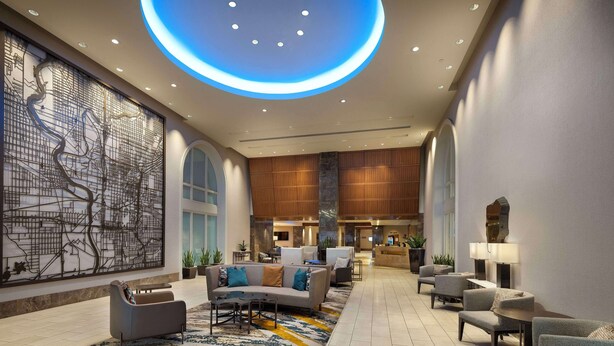 A spacious lobby with a modern design, featuring a large seating area with sofas and armchairs, a decorative wall with a cityscape mural, and a circular ceiling light. - Hilton Indianapolis Hotel & Suites (Indianapolis)
