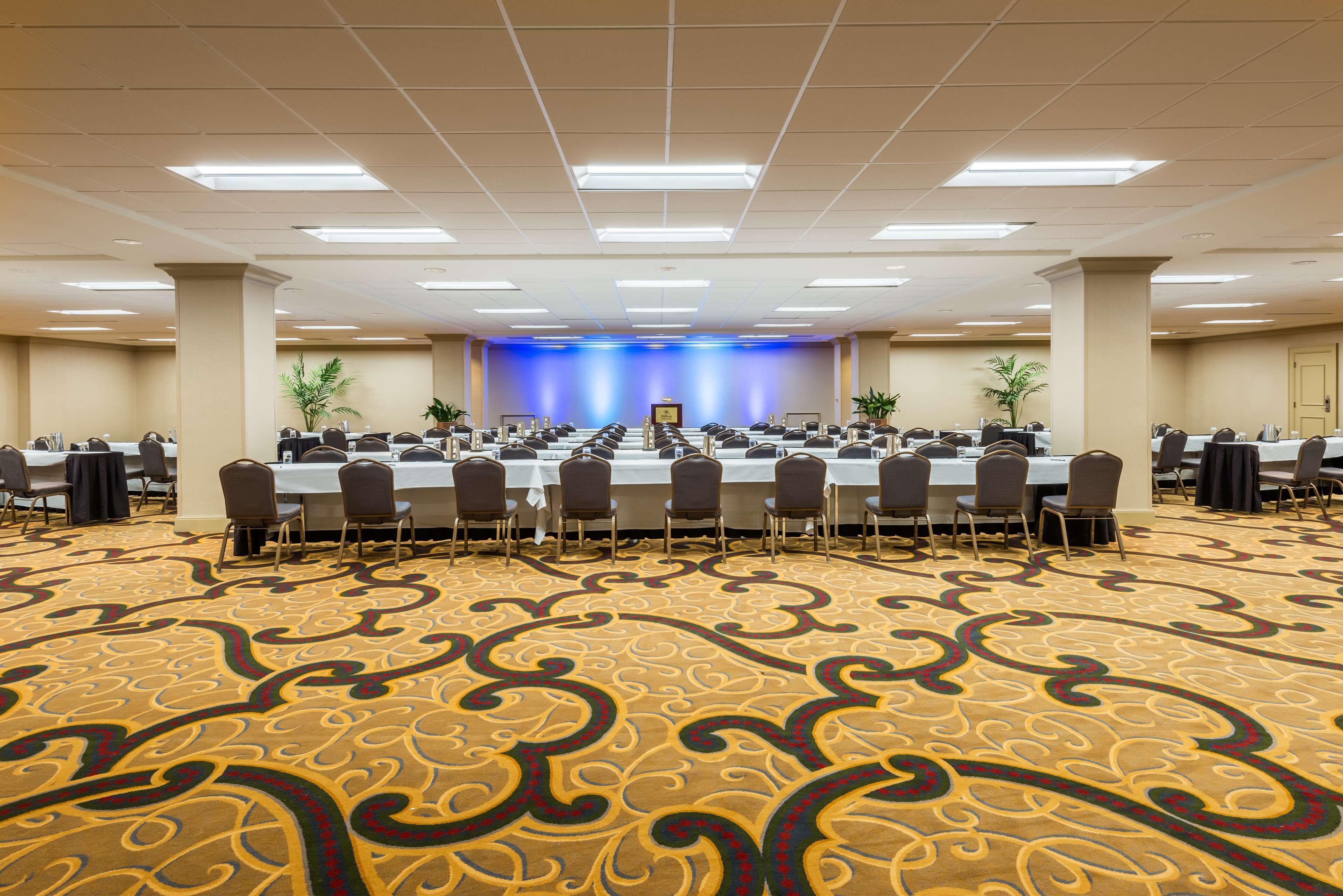 Un salón de conferencias con una larga mesa, varias sillas y un escenario con un fondo azul.