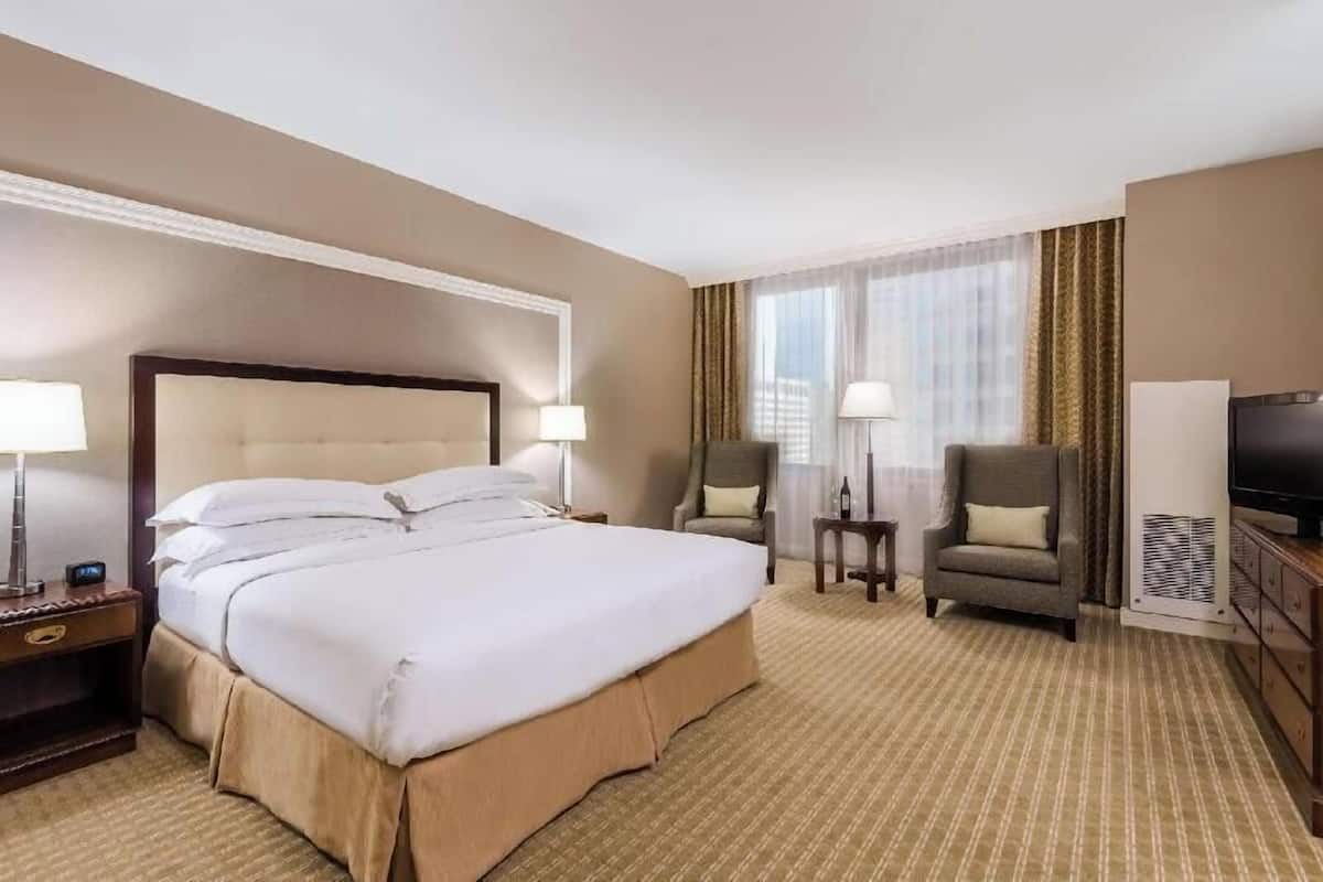 Quarto de hotel com uma cama grande, duas cadeiras, uma TV e uma mesinha.