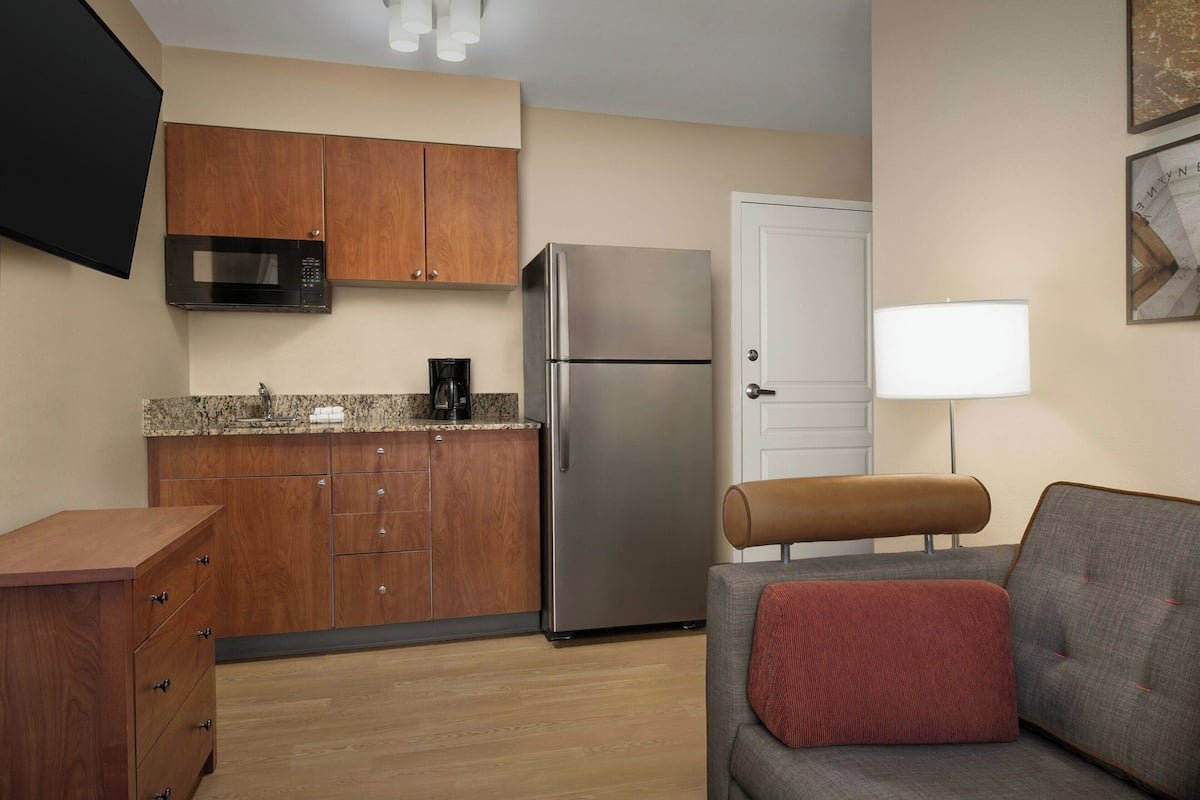 A compact kitchen with wooden cabinets, a stainless steel refrigerator, a microwave, and a coffee maker. There is a gray sofa with a red pillow and a white door in the background.