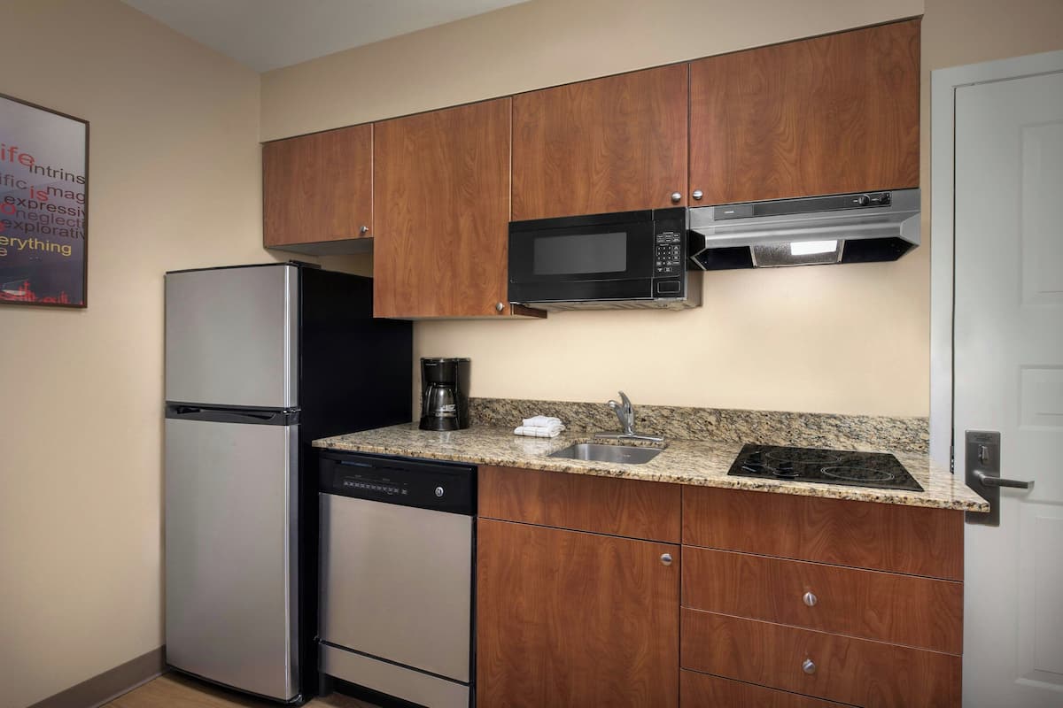 A compact kitchen with wooden cabinets, a stainless steel refrigerator, a microwave, a dishwasher, and a granite countertop.