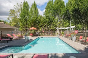 A rectangular swimming pool with a tiled edge, surrounded by a metal fence. There are lounge chairs and a small table with a red umbrella. - TownePlace Suites by Marriott Portland Hillsboro (Hillsboro)