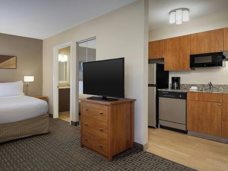 A hotel room with a bed, a television on a wooden dresser, a kitchen with a microwave, a coffee maker, and a dishwasher.