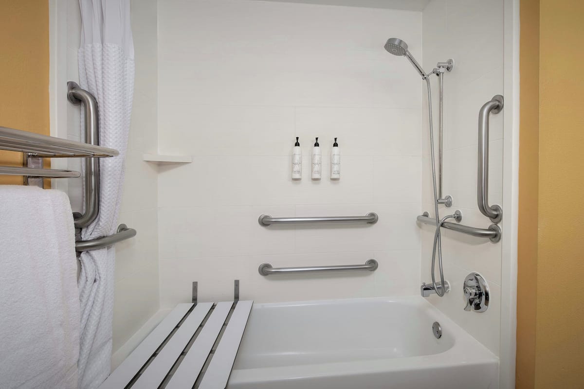 A bathroom with a white bathtub, grab bars, a showerhead, and three bottles of shampoo.