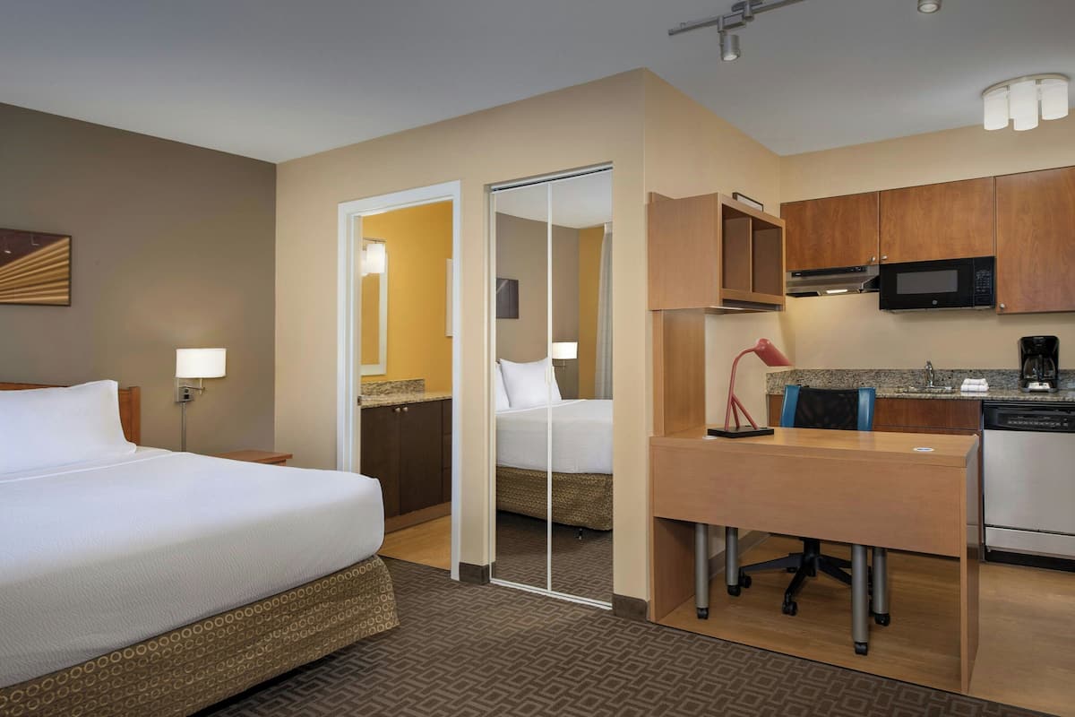A hotel room with a bed, a desk, a kitchenette, and a bathroom.