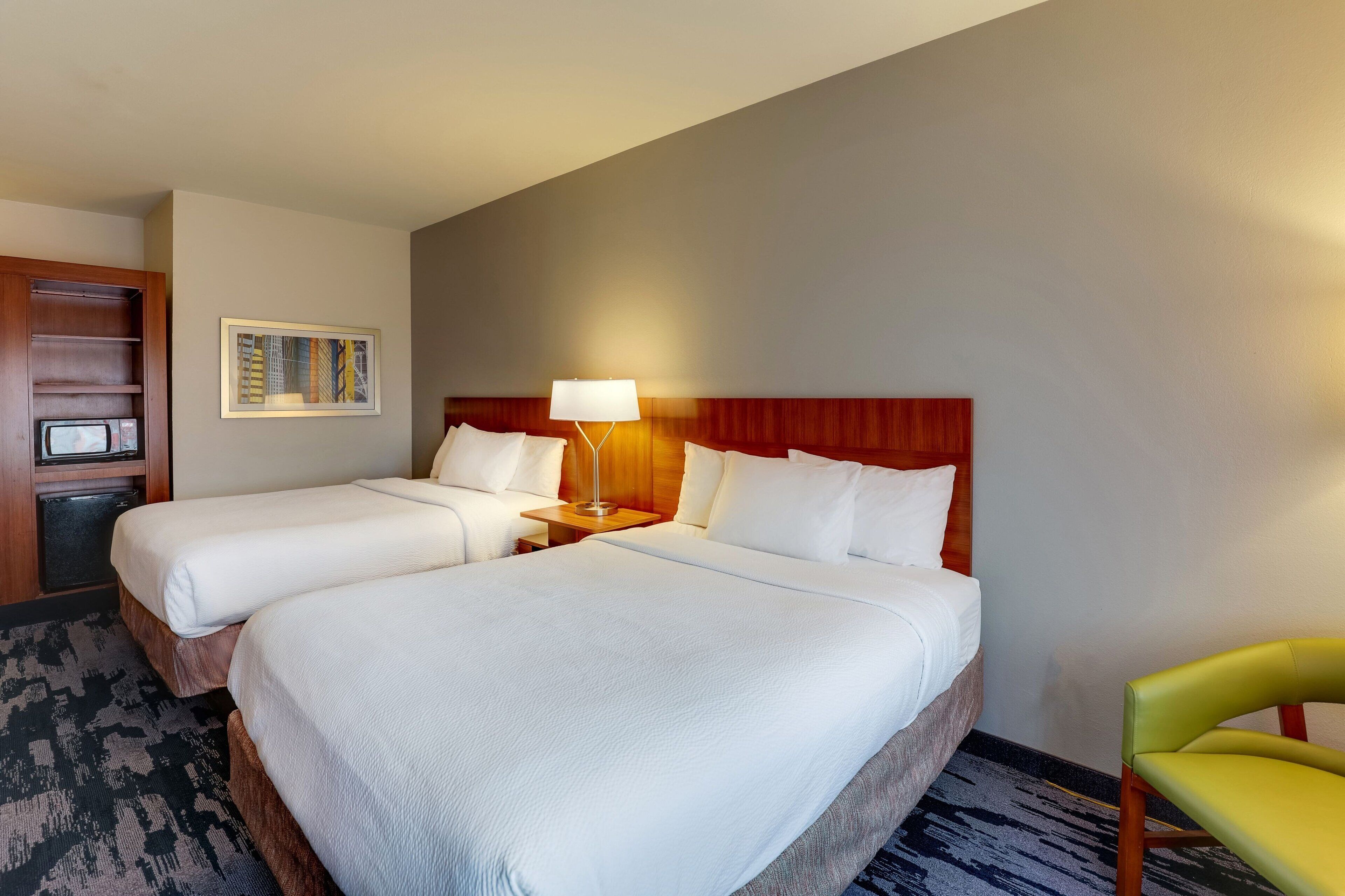 Une chambre d’hôtel avec deux lits, une tête de lit en bois, une table de chevet avec une lampe, un tableau encadré au mur et un fauteuil vert.