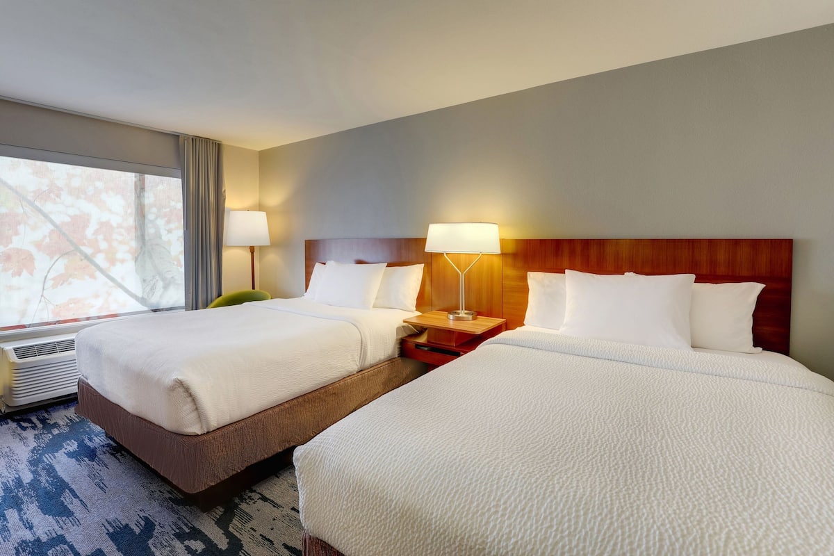 Ein Hotelzimmer mit zwei Betten, einem hölzernen Kopfteil, Nachttischlampen und einem großen Fenster mit Vorhängen.