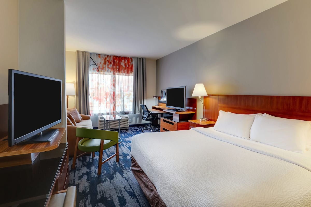 Ein Hotelzimmer mit einem großen Bett, einem Schreibtisch mit Fernseher, einem Sessel, einer Schreibtischlampe und einem Fenster mit Vorhängen.