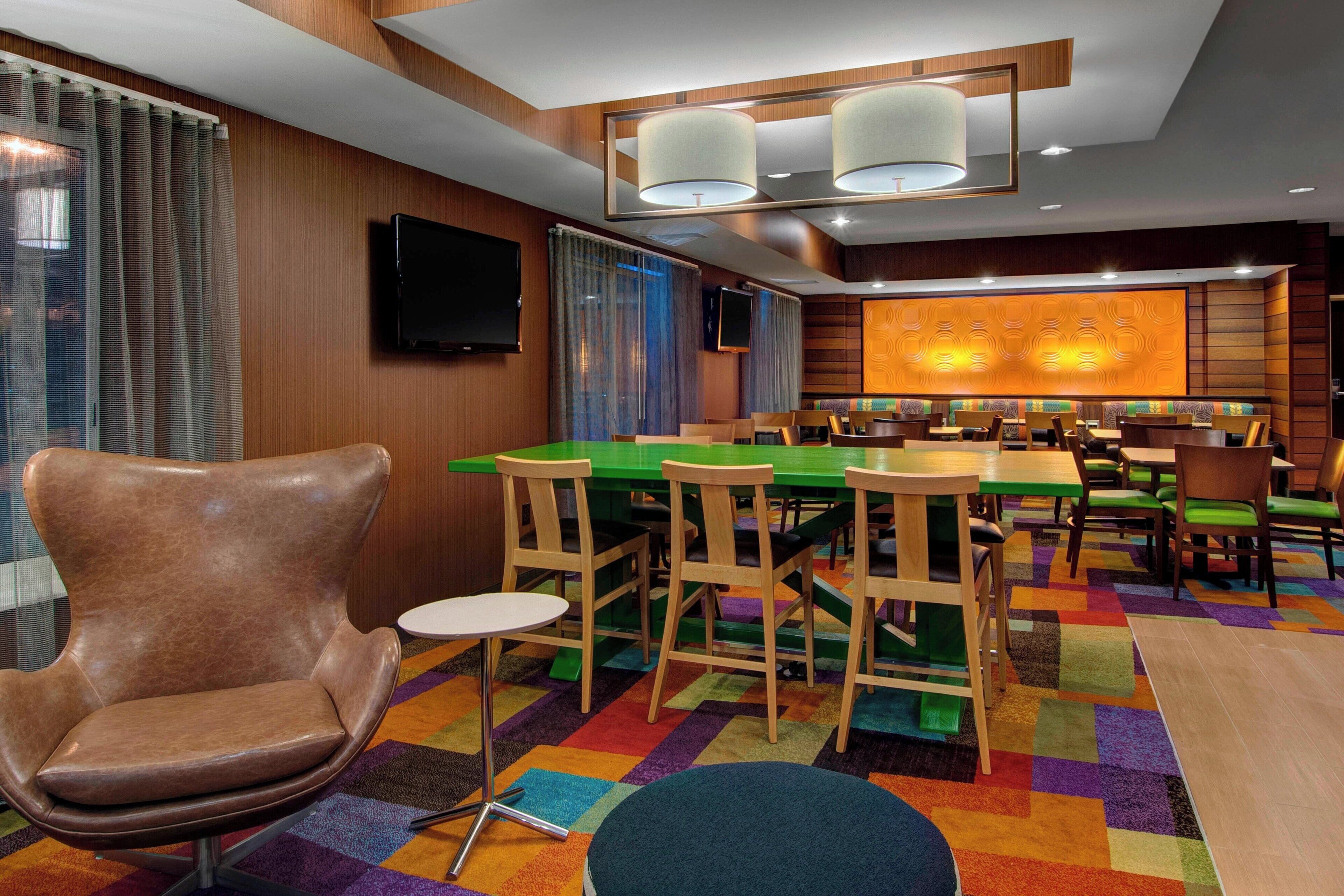 A hotel room with a dining area featuring a green table, wooden chairs, and a colorful carpet.