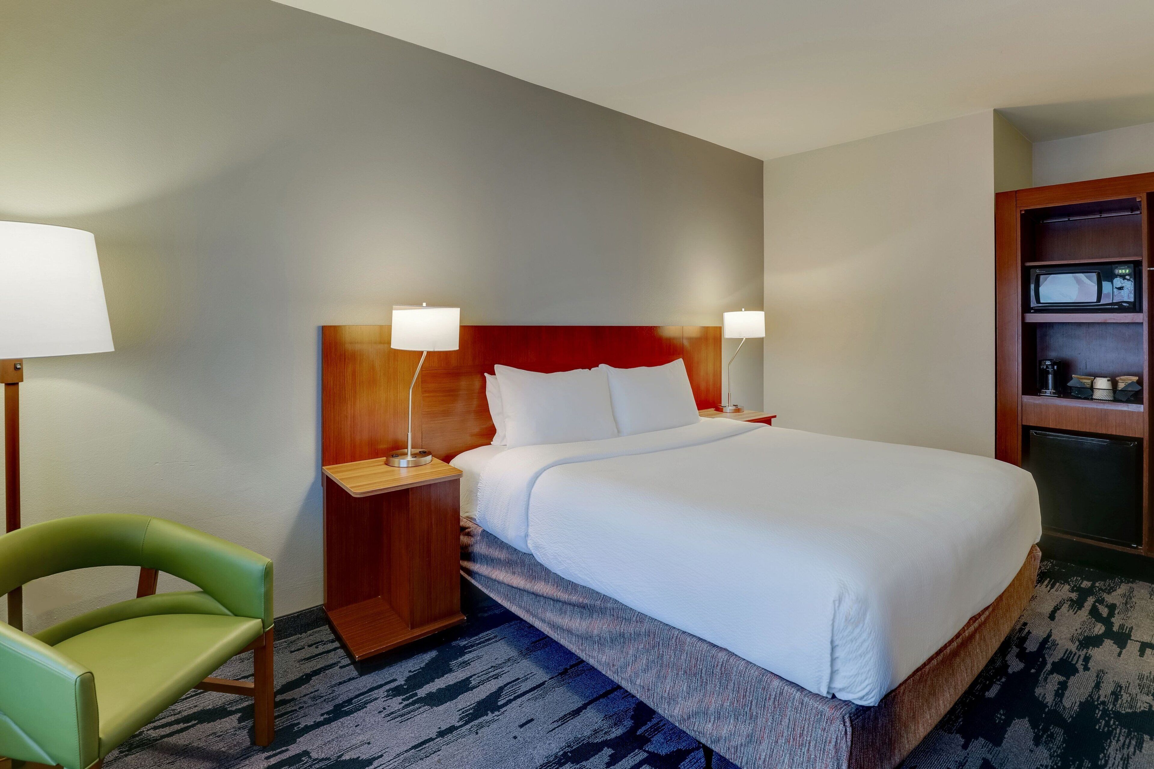 A hotel room with a large bed, two bedside tables with lamps, a green chair, and a wooden cabinet with a microwave.