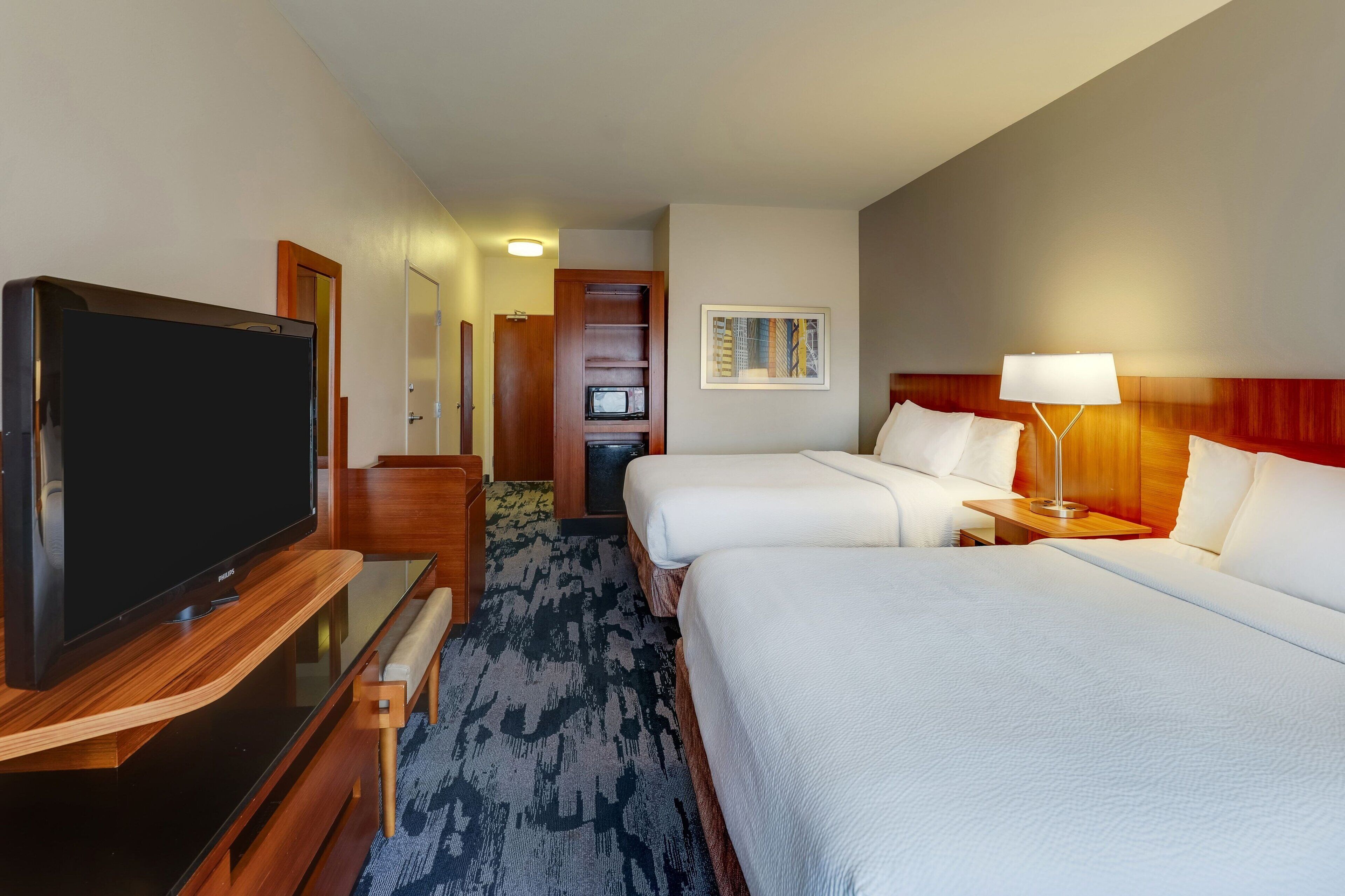 Ein Hotelzimmer mit Flachbildfernseher, zwei Betten, einem Schreibtisch mit Stuhl, einer Lampe und einem gerahmten Bild an der Wand.