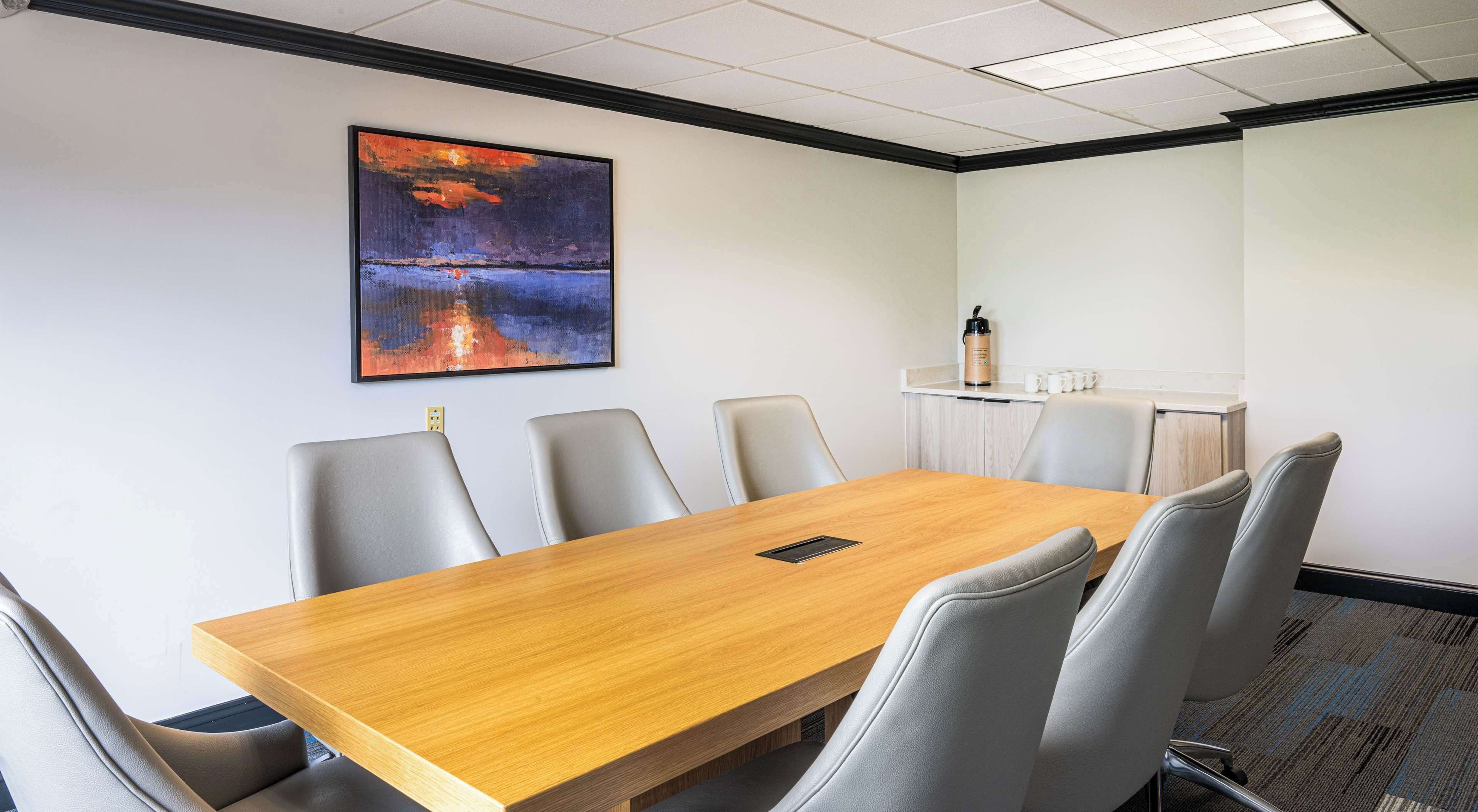 Un salón de conferencias con una mesa de madera, seis sillas grises, un cuadro de un atardecer y una pizarra.