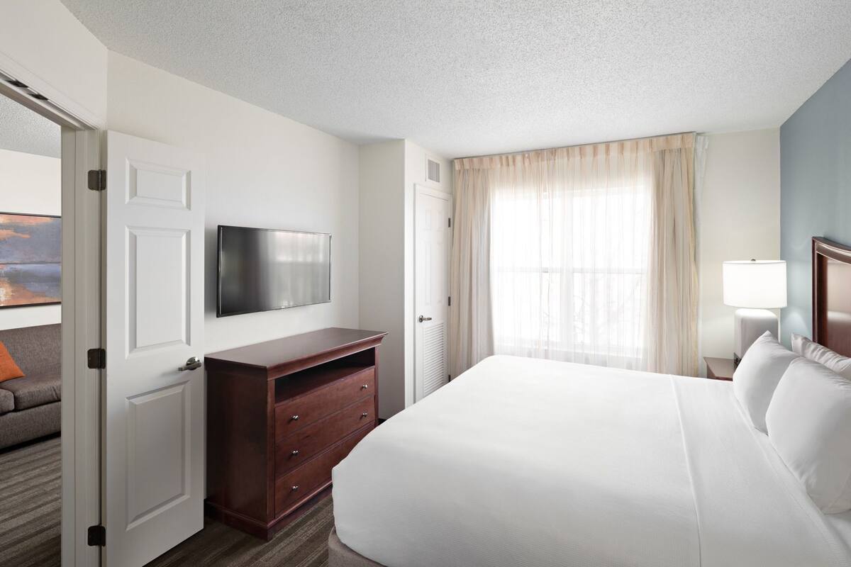 A hotel room with a bed, a TV mounted on the wall, a wooden dresser, a window with curtains, and a nightstand with a lamp.