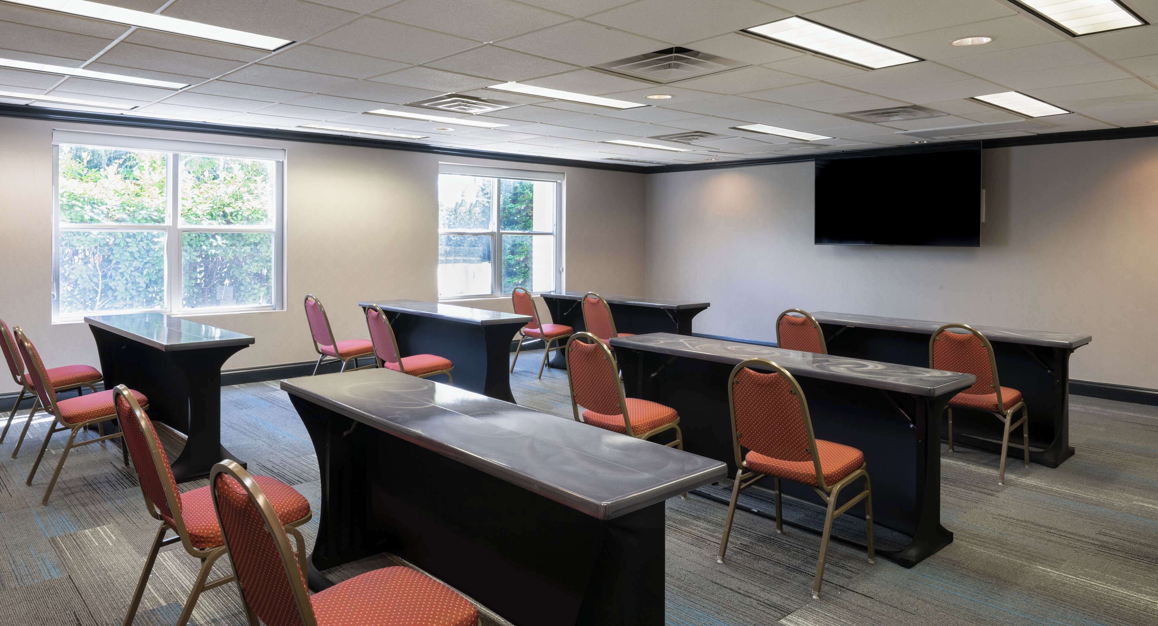 A conference room with multiple tables and chairs, large windows, and a mounted screen on the wall.