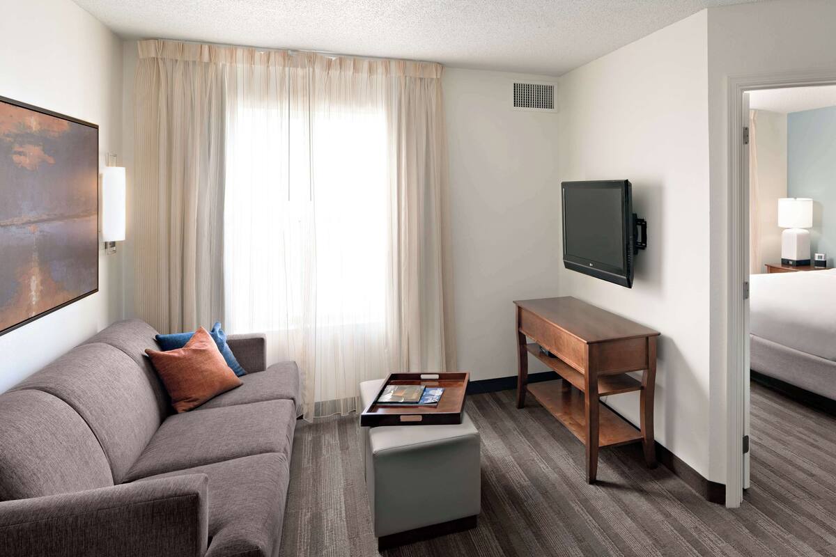 A hotel room with a grey sofa, a small wooden table, a flat-screen TV, and a large window with curtains.