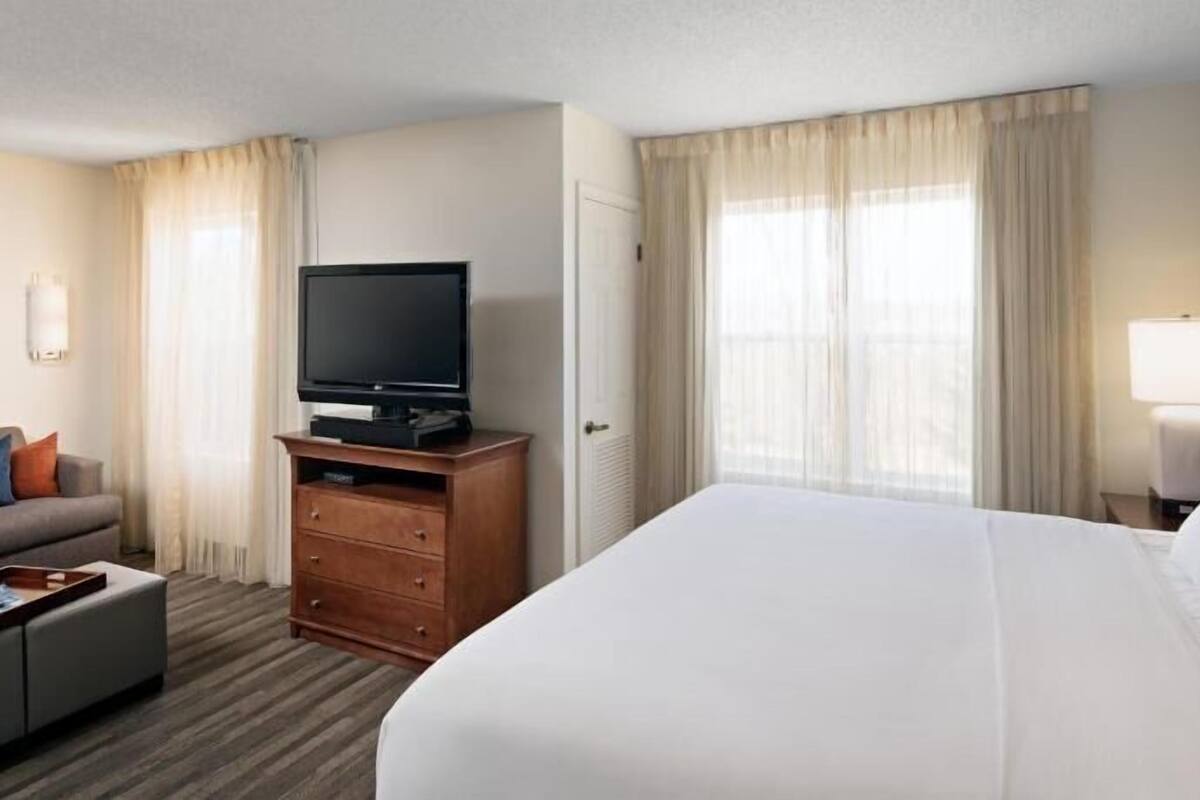 A hotel room with a bed, a TV on a wooden stand, a desk with a lamp, and a window with curtains.