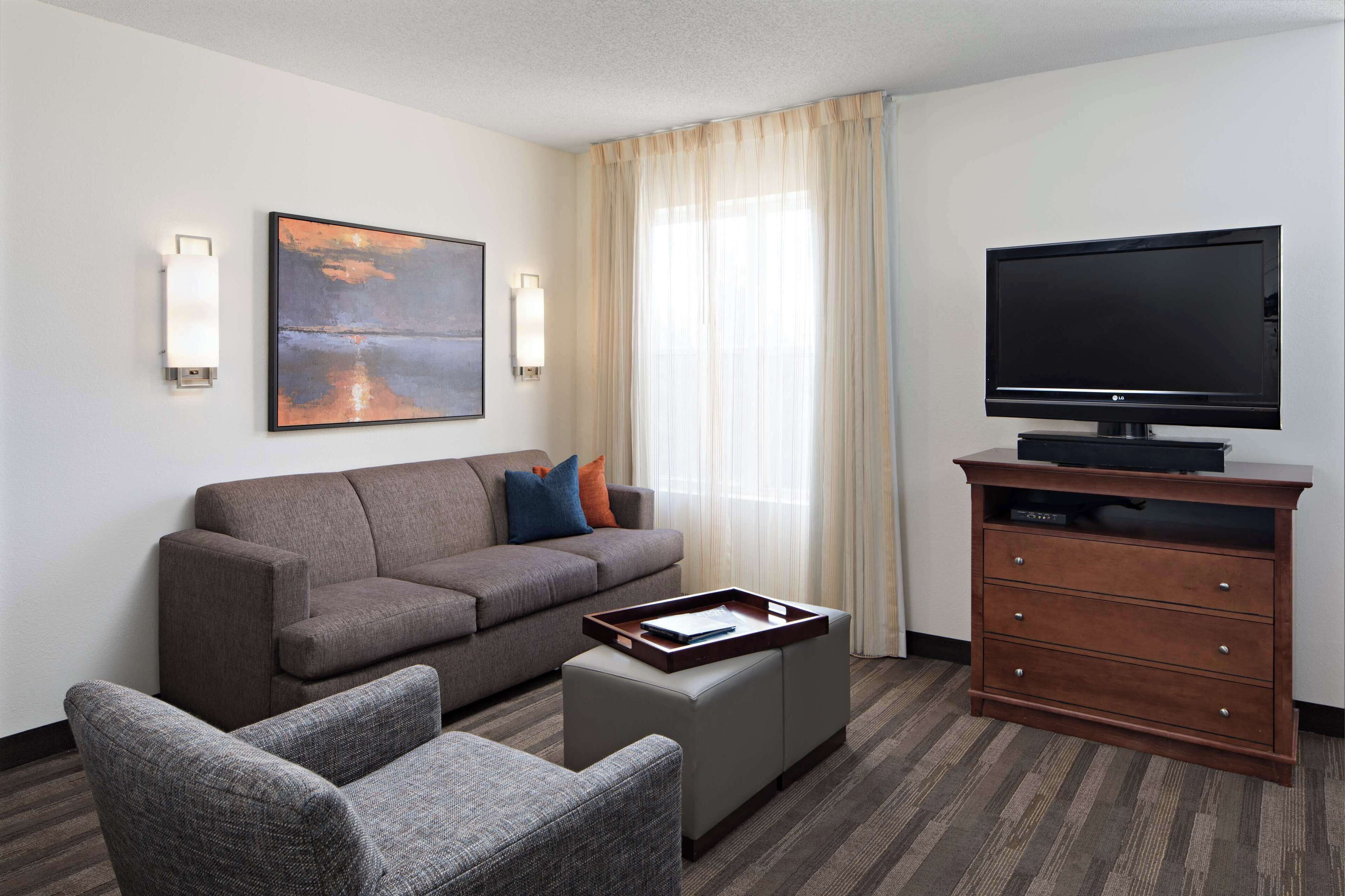 Une chambre d’hôtel avec un canapé gris, un fauteuil gris, un meuble TV en bois et un tableau accroché au mur.