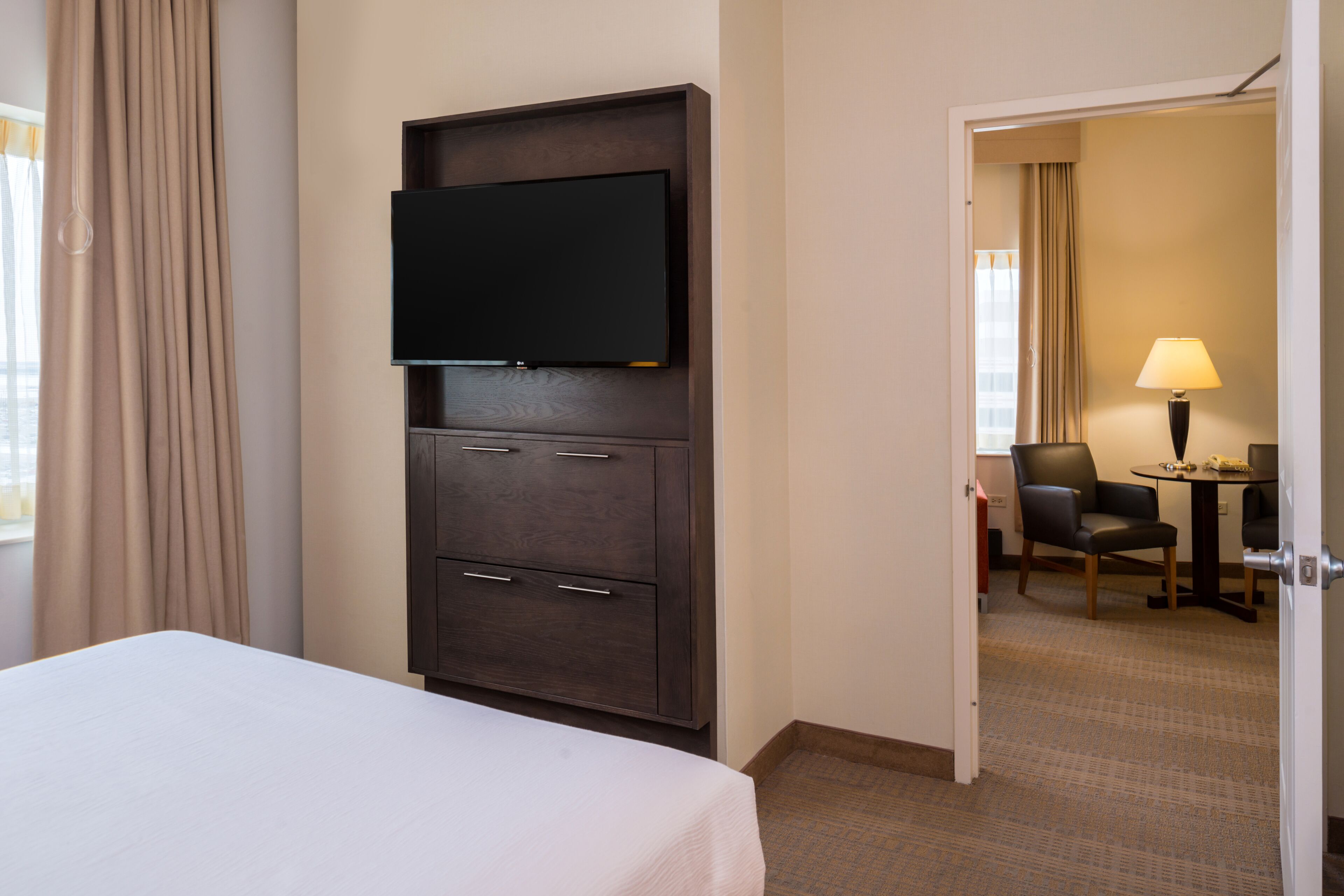 A hotel room with a bed, a dark wooden cabinet with a flat-screen TV mounted above it, a desk with a chair, and a lamp.