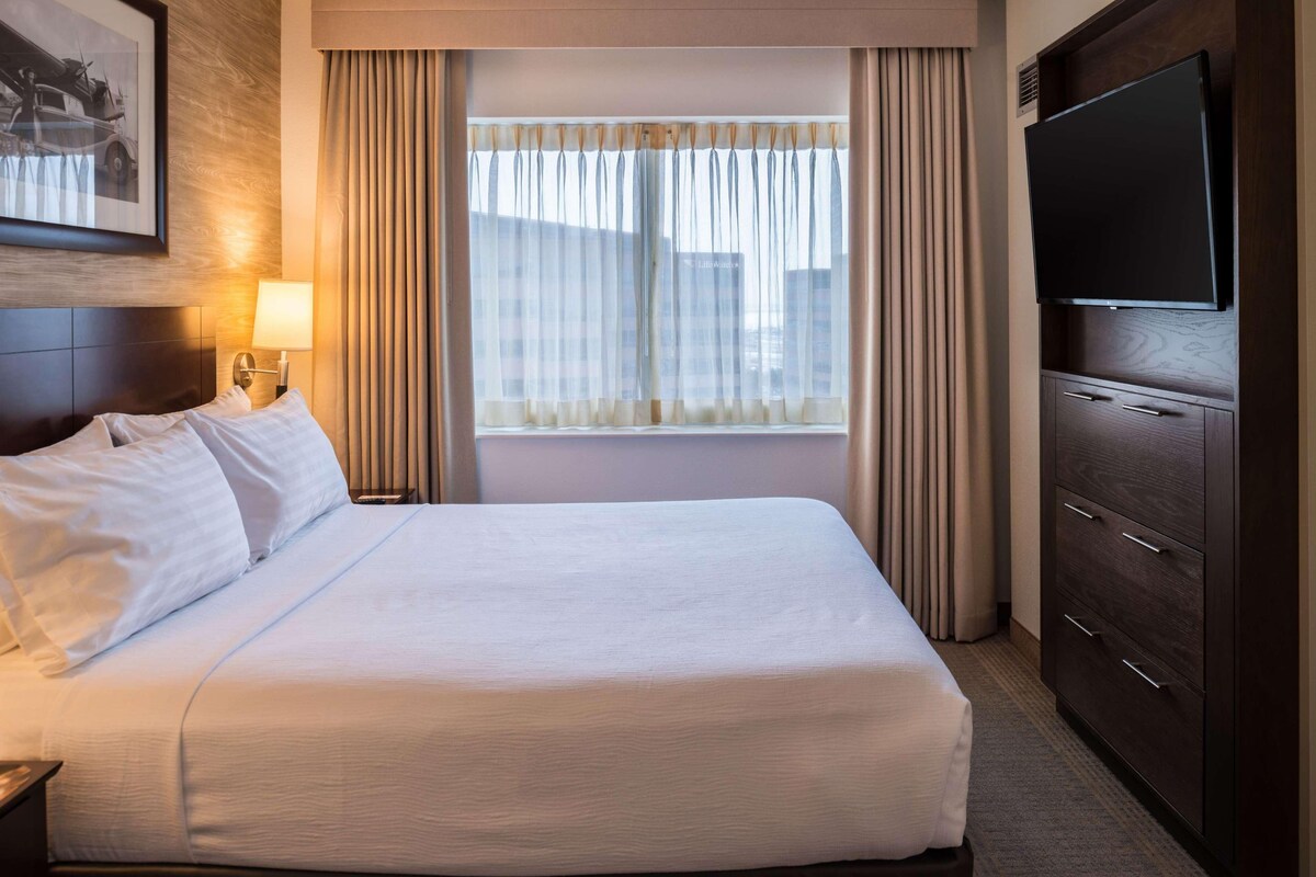 Una habitación de hotel con cama, televisión, lámpara y ventana con cortinas.