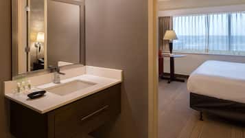 A hotel room with a large mirror, a sink, a bedside table with a lamp, a bed, and a view of the sea through the window.