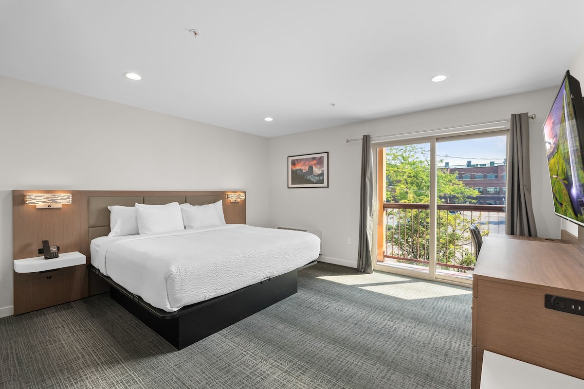 A modern hotel room with a large bed, a desk, a TV, and a window with a city view.