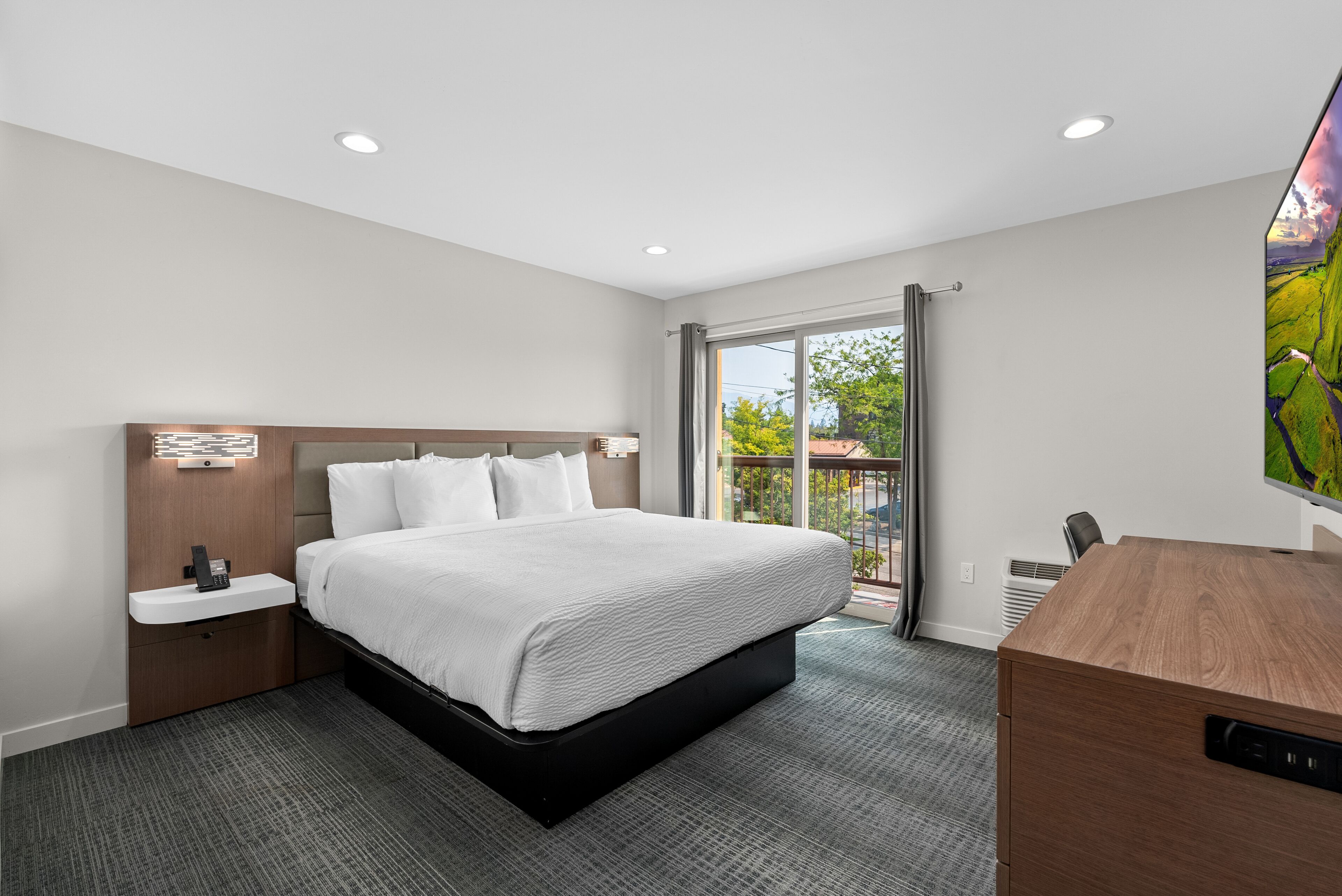 A modern hotel room with a large bed, a bedside table with a phone, a desk with a chair, and a large window with a view of greenery.