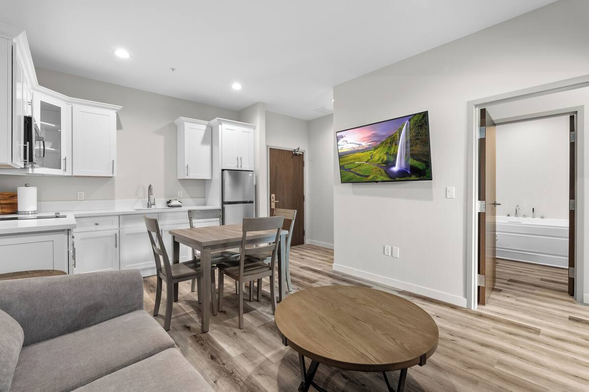 A modern kitchen with white cabinets, a dining area with a wooden table and chairs, and a large TV mounted on the wall.