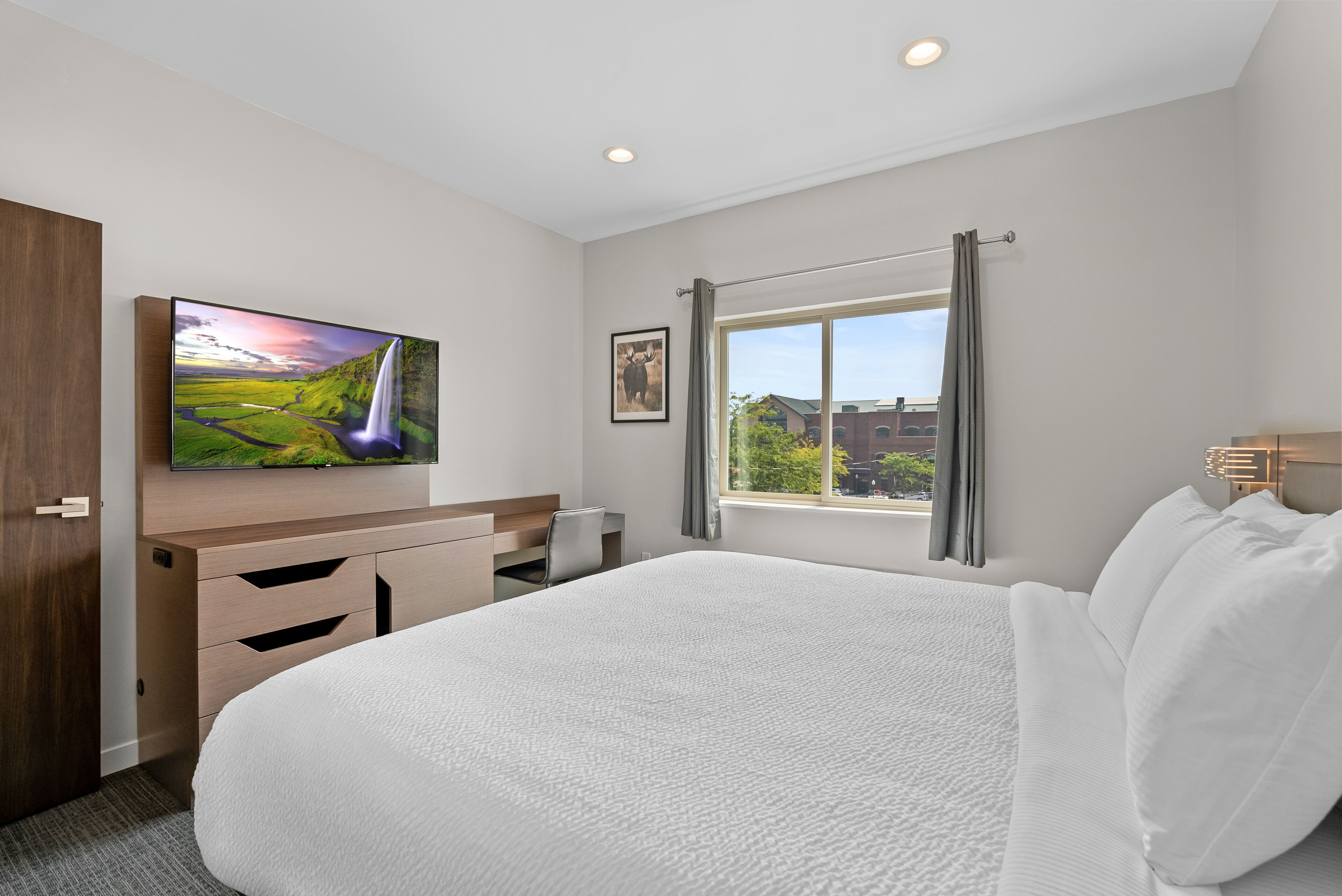 A hotel room with a large bed, a desk with a chair, a flat-screen TV mounted on the wall, a window with curtains, and a view of buildings outside.