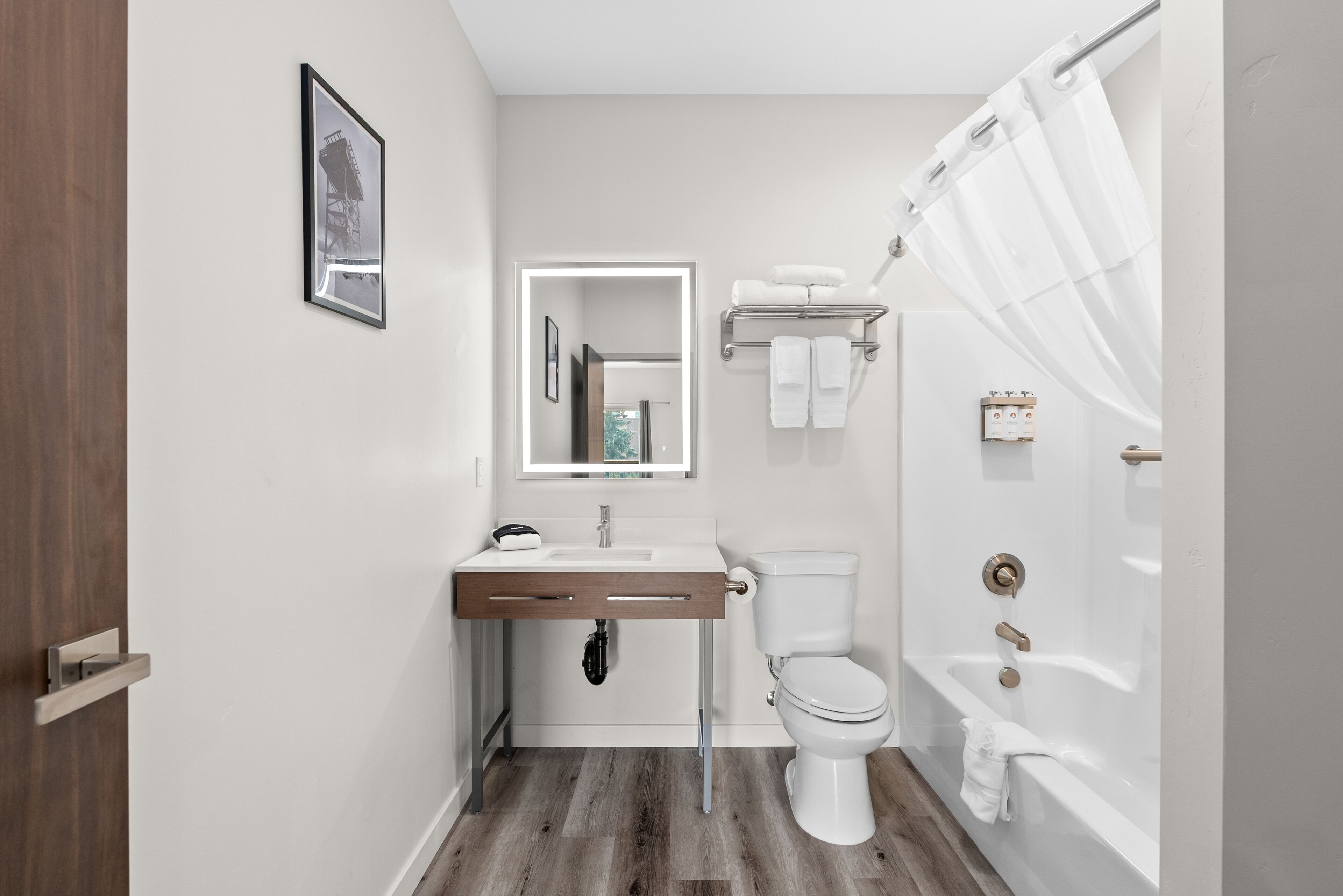 A bathroom with a wooden floor, white fixtures, a shower curtain, and a framed picture on the wall.