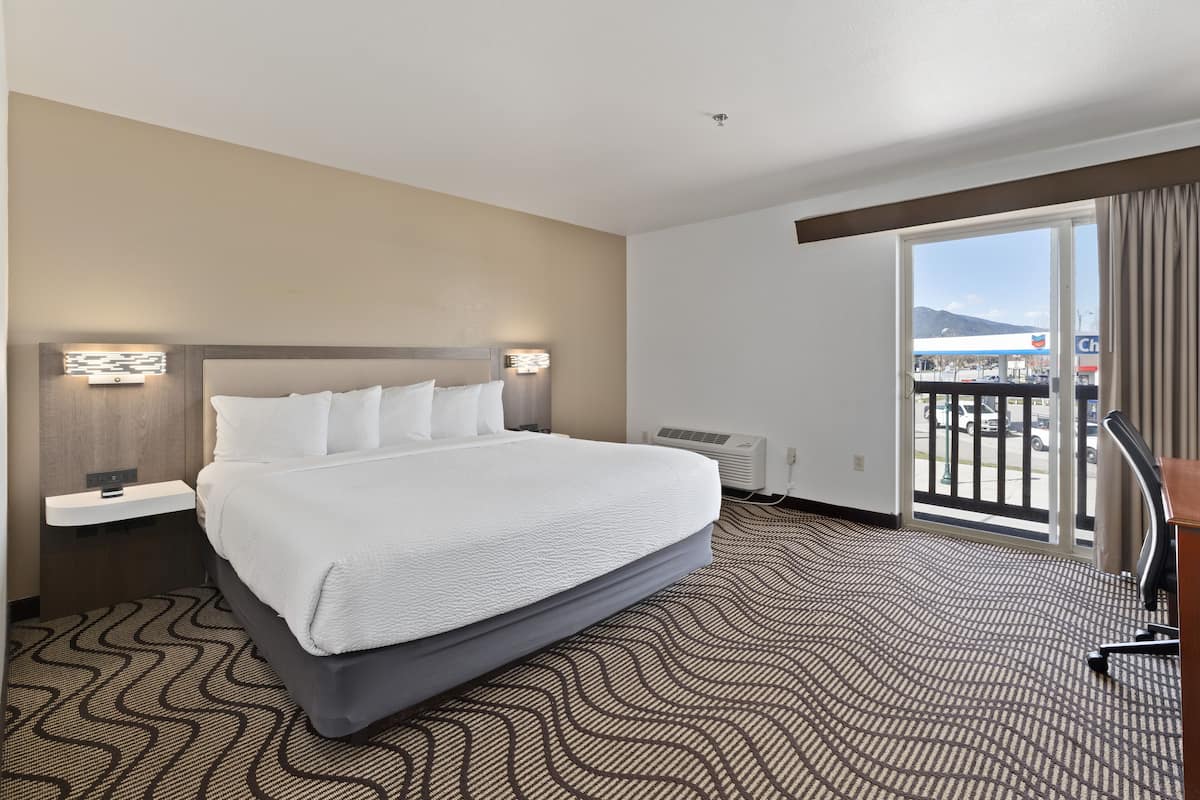 A hotel room with a large bed, a desk, a chair, a window with a view of a parking lot and mountains, and a patterned carpet.