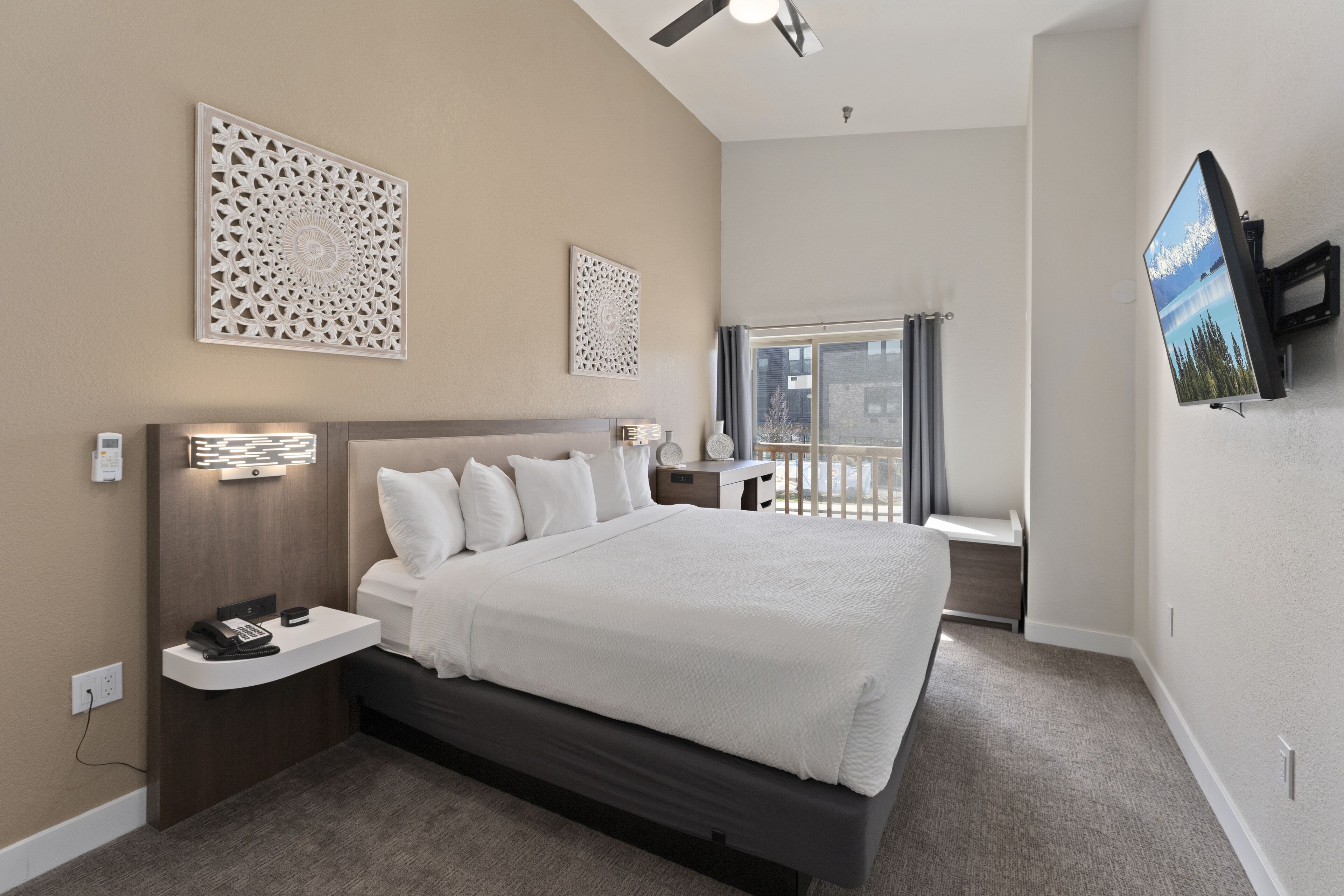 A modern hotel room with a large bed, a desk with a phone, decorative wall art, a TV, and a window with a view.