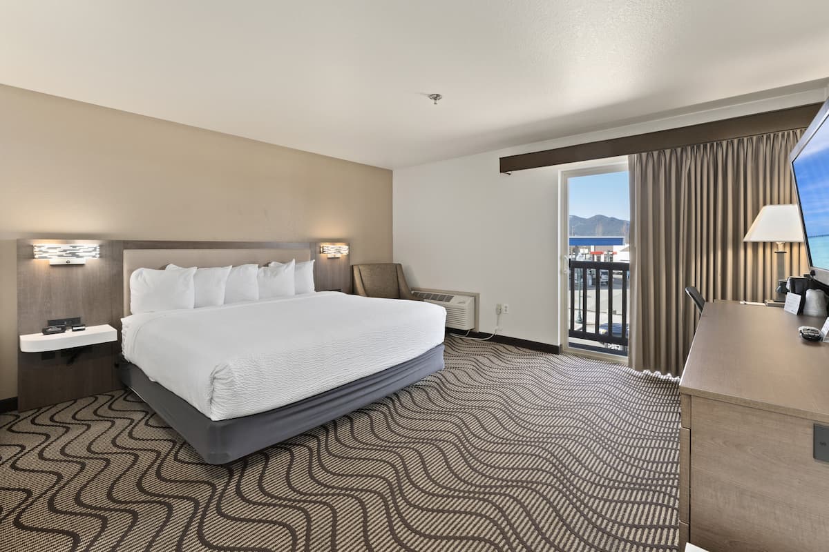 A hotel room with a large bed, a desk, a chair, a nightstand, a lamp, and a view of the sea through a window.