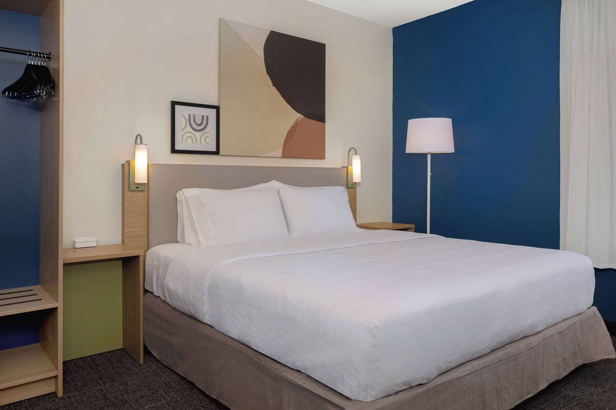 A neatly made bed with a white comforter and pillows, a bedside table with a lamp, a wall-mounted abstract painting, and a hanging coat rack.