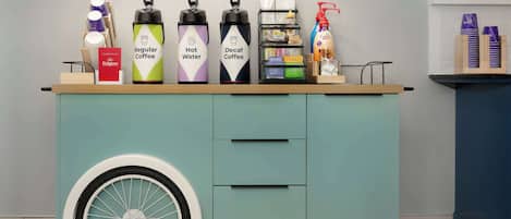 Un espace dédié aux boissons, équipé de distributeurs étiquetés pour le café ordinaire, le café chaud et le café décaféiné, ainsi que d’un distributeur de lait et d’une bouteille de jus.