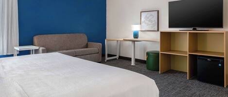 A hotel room with a large bed, a desk with a lamp, a television, and a small refrigerator.