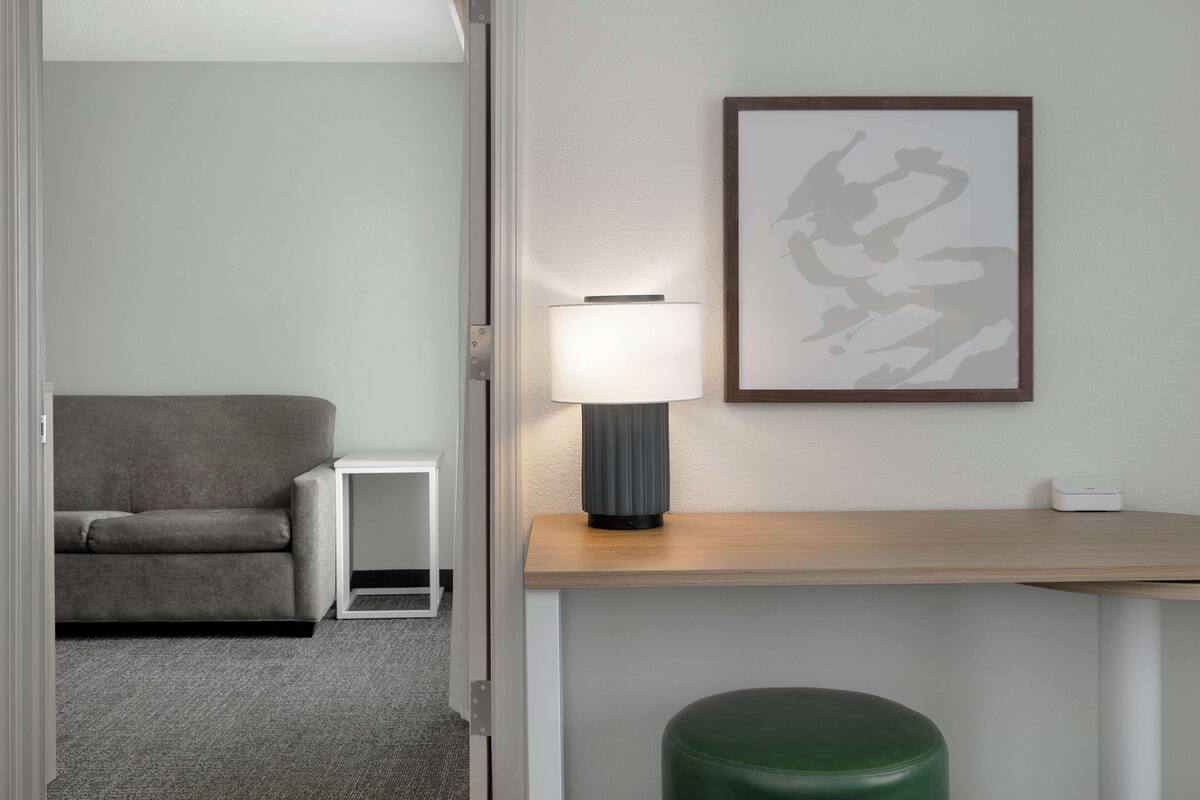 A modern room with a grey sofa, a small white side table, a green ottoman, a wooden desk, a framed abstract artwork, and a floor lamp.