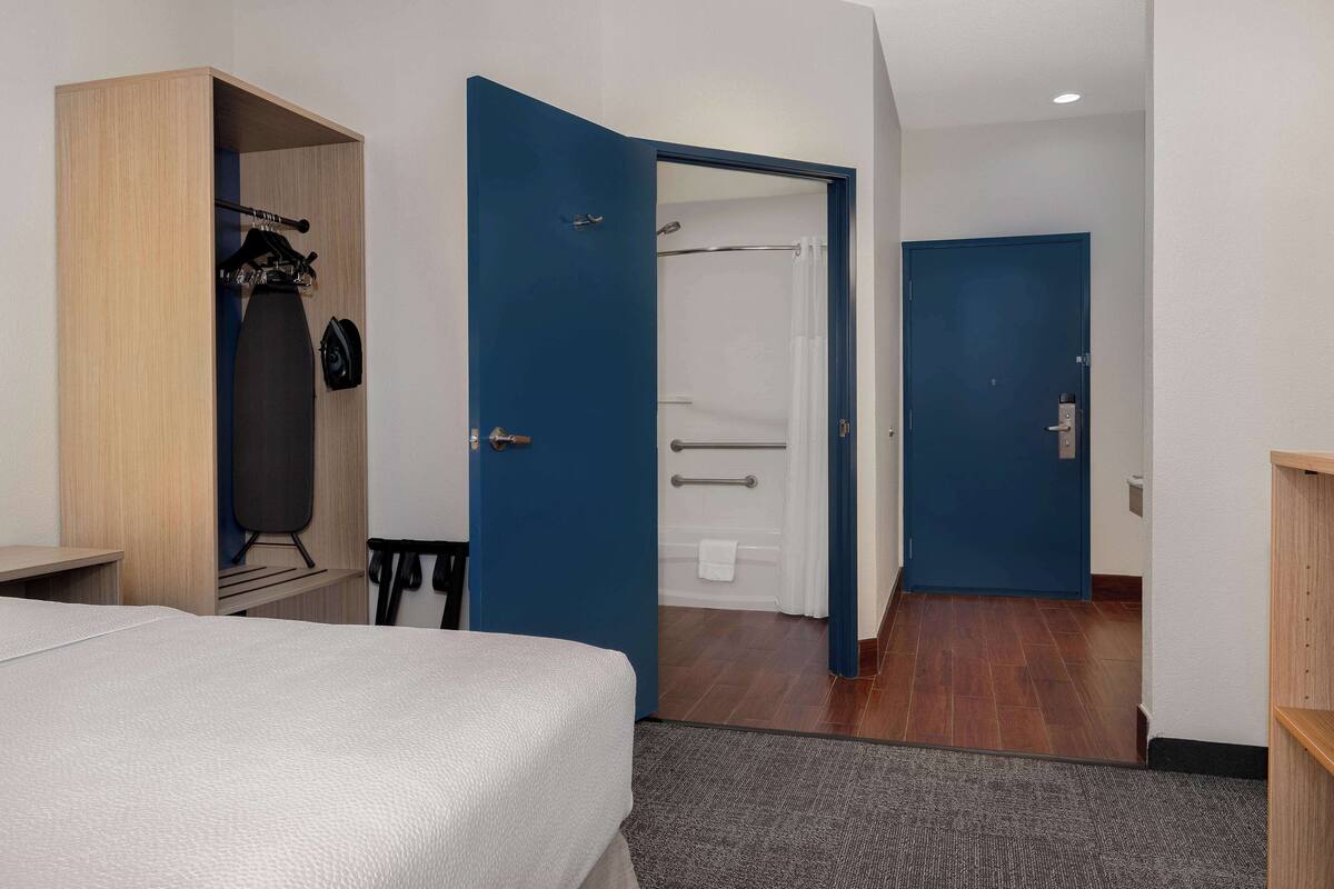 A hotel room with a bed, a wardrobe, a bathroom with a shower, and a door leading to another room.